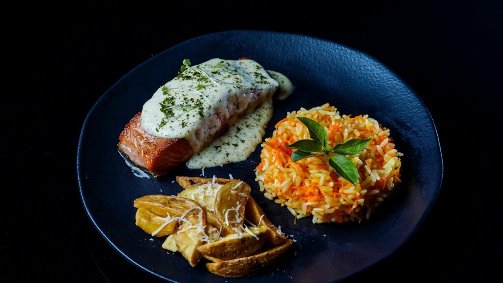 Delicious salmon served with creamy sauce, rice pilaf, and potato wedges on a black plate.
